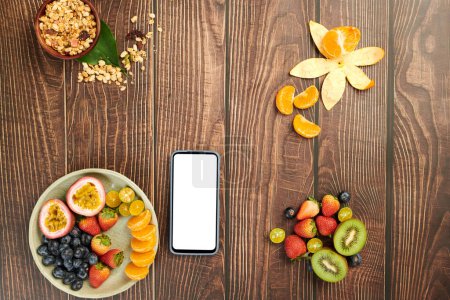 Foto de Smartphone con pantalla en blanco en mesa de madera con frutas y granola cuenco servido para el desayuno - Imagen libre de derechos