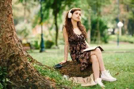 Téléchargez les photos : Jeune femme pensive assise sous un arbre, écoutant de la musique et écrivant dans un journal - en image libre de droit