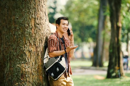 Téléchargez les photos : Souriant jeune homme appuyé sur l'arbre en parlant au téléphone avec un ami - en image libre de droit