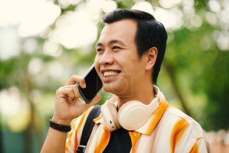 Téléchargez les photos : Portrait de jeune Vietnamien souriant parlant au téléphone avec un ami en marchant à l'extérieur - en image libre de droit