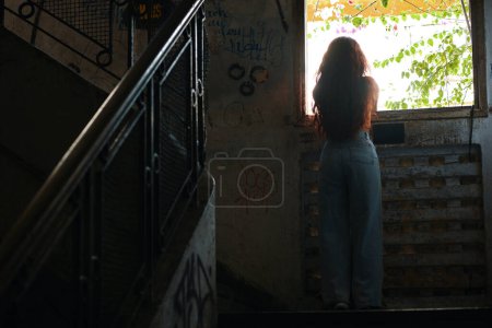 Téléchargez les photos : Jeune femme aux cheveux longs regardant par fenêtre de bâtiment abandonné et en décomposition - en image libre de droit