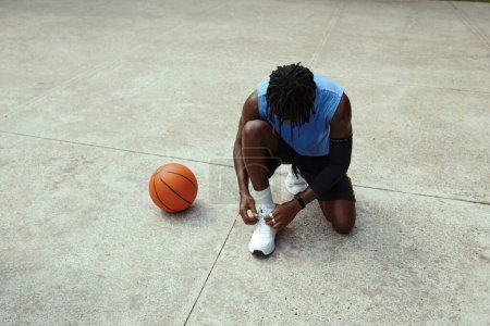 Foto de Jugador de streetball negro atando cordones de zapatos cuando espera a un amigo en la cancha - Imagen libre de derechos