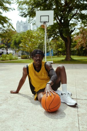 Téléchargez les photos : Joueur de streetball joyeux assis sur le sol à côté de la balle se reposant après le match - en image libre de droit