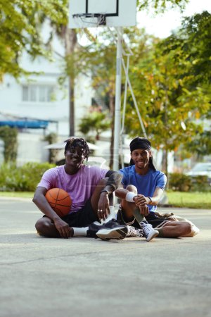 Téléchargez les photos : Joueurs de streetball noirs souriants assis sur le terrain du court extérieur - en image libre de droit