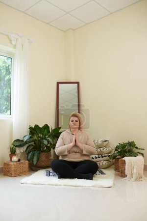 Téléchargez les photos : Femme calme et détendue croisant les jambes tout en étant assise sur un tapis en position lotus pendant la méditation - en image libre de droit