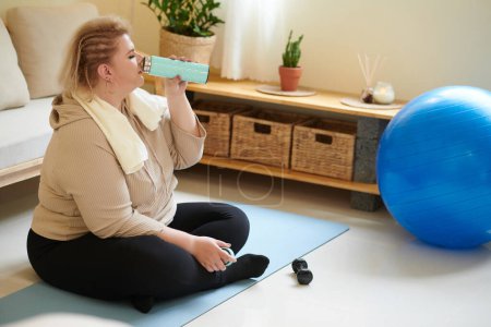 Foto de Mujer joven de tamaño grande sentada en una esterilla de yoga y bebiendo agua dulce después de entrenar en casa - Imagen libre de derechos