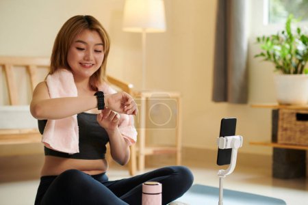 Photo for Smiling sweaty young Asian woman checking smartwatch after personal online training - Royalty Free Image