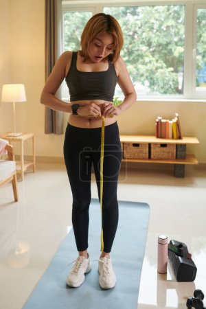 Téléchargez les photos : Jeune femme excitée mesurant la taille après l'exercice à la maison - en image libre de droit