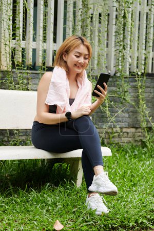 Photo for Fit young Asian sportswoman sitting on bench in park and video calling friend after outdoor training - Royalty Free Image