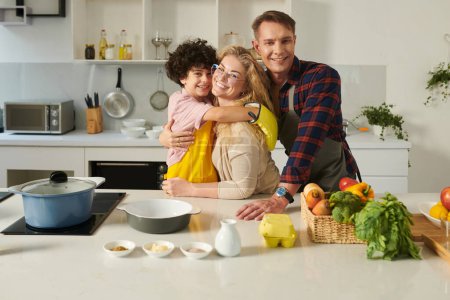 Foto de Familia feliz de tres de pie en el mostrador de la cocina, abrazando y mirando a la cámara - Imagen libre de derechos