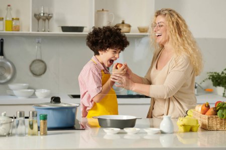 Photo for Funny boy trying to bite apple that mother prepared for baking pie - Royalty Free Image