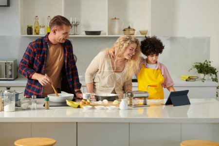 Téléchargez les photos : Famille heureuse suivant la recette sur tablette lors de la cuisson des pâtes à la maison - en image libre de droit