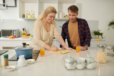 Téléchargez les photos : Couple souriant vérifier les mesures pour la recette de plat écrit dans le carnet - en image libre de droit