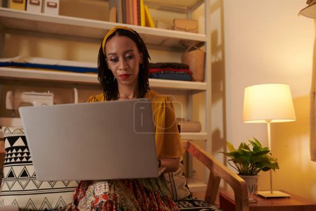 Téléchargez les photos : Femme couturière sérieuse travaillant sur ordinateur portable tard dans la nuit dans son bureau à la maison - en image libre de droit