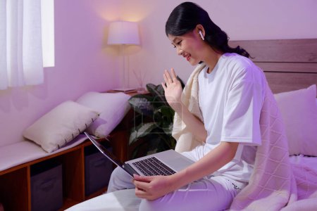 Téléchargez les photos : Joyeux jeune femme assise sur le lit dans la lumière au néon et appel vidéo ami - en image libre de droit