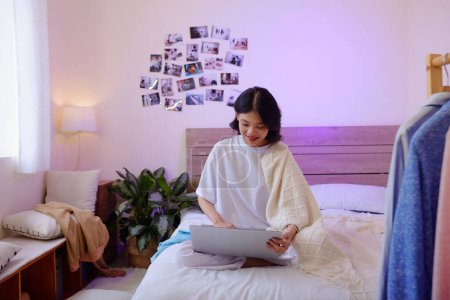 Téléchargez les photos : Souriante fille assise sur le lit sous la couverture de lancer et de travailler sur ordinateur portable - en image libre de droit