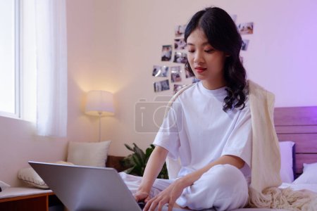 Téléchargez les photos : Adolescente en pyjama de coton travaillant sur un ordinateur portable lorsqu'elle est assise sur le lit à la maison - en image libre de droit