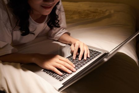 Foto de Imagen recortada de una adolescente sonriente trabajando en una computadora portátil hasta altas horas de la noche cuando está acostada en la cama - Imagen libre de derechos