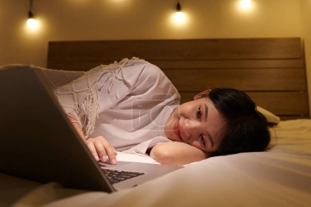 Téléchargez les photos : Souriante fille reposant sur le lit et regardant film comique romantique sur ordinateur portable - en image libre de droit