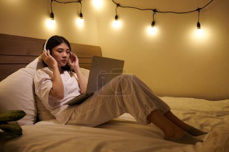 Foto de Adolescente en ropa de salón en los auriculares viendo webinar en el ordenador portátil al acostarse en la cama en casa - Imagen libre de derechos