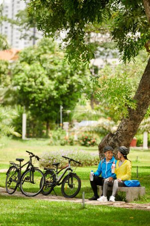 Téléchargez les photos : Heureux jeune couple vietnamien boire de l'eau douce sous l'arbre dans le parc après une balade à vélo - en image libre de droit