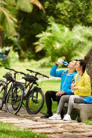 Téléchargez les photos : Soif jeune couple boire de l'eau lorsque vous vous reposez sur le banc après une balade à vélo - en image libre de droit