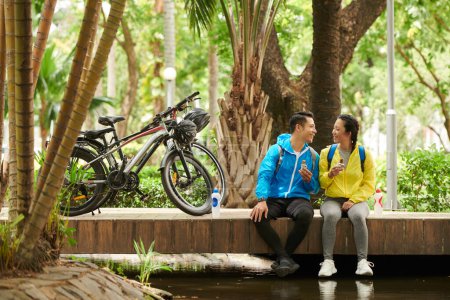 Téléchargez les photos : Couple heureux en amour manger des barres d'énergie granola pour grignoter lorsque vous vous reposez sur le pont après une balade à vélo - en image libre de droit