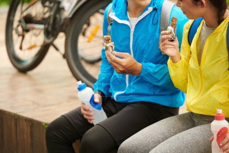 Téléchargez les photos : Image recadrée de jeunes couples mangeant des barres de granola pour obtenir un peu d'énergie pour la randonnée à vélo - en image libre de droit