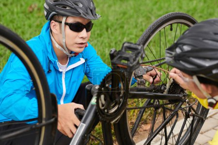 Téléchargez les photos : Homme sérieux dans le casque et les lunettes de soleil vérifiant si la roue du vélo est bien ajustée et le levier de déverrouillage rapide est sécurisé - en image libre de droit