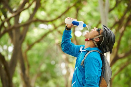 Photo for Thirsty cyclist in windbreaker, helmet and sunglasses drinking fresh water - Royalty Free Image