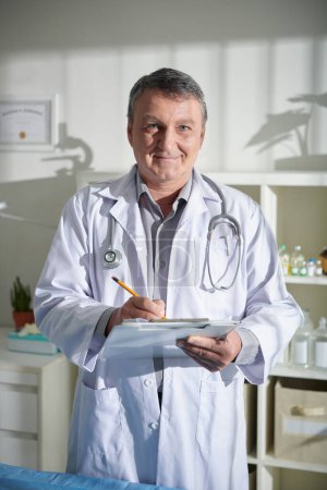 Foto de Retrato del veterinario maduro positivo que llena el formulario médico en el portapapeles - Imagen libre de derechos