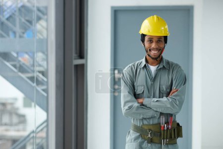 Téléchargez les photos : Portrait du contremaître noir positif en uniforme et casque de sécurité debout à l'intérieur du bâtiment en construction - en image libre de droit