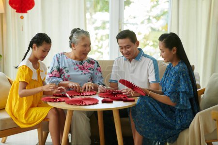 Téléchargez les photos : Famille de trois générations fabriquant des éventails en papier rouge pour décorer la maison pour Tet - en image libre de droit