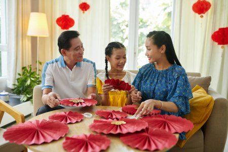 Foto de Padres e hija haciendo abanicos tradicionales de papel rojo para decorar el apartamento para la celebración del Tet - Imagen libre de derechos
