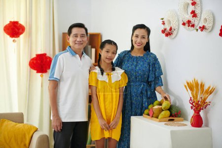 Foto de Los padres sonrientes y la hija adolescente de pie a la mesa con frutas preparadas para la celebración del Tet - Imagen libre de derechos