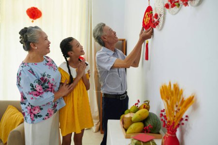 Téléchargez les photos : Grands-parents et petite-fille décorant les murs de l'appartement avec des ornements traditionnels - en image libre de droit