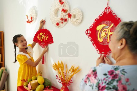 Foto de Sonriente chica vietnamita ayudando a la abuela a decorar las paredes del apartamento para la celebración del Tet - Imagen libre de derechos