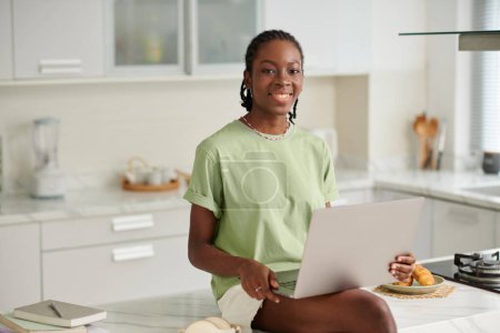 Téléchargez les photos : Portrait de joyeuse fille noire avec ordinateur portable assis sur l'îlot de cuisine - en image libre de droit