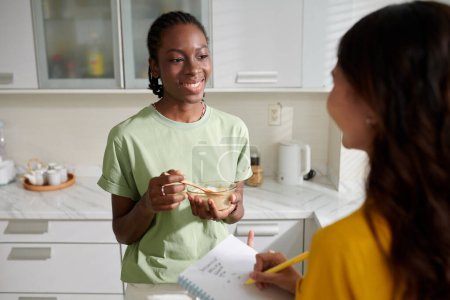 Photo for Smiling girl eating granola and talking to roommate in kitchen - Royalty Free Image