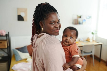 Téléchargez les photos : Portrait de femme noire avec petit bébé dans les mains debout dans la chambre - en image libre de droit