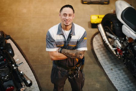 Photo for Portrait of happy tattooed motorcycle mechanic crossing arms and smiling at camera - Royalty Free Image