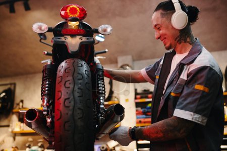 Foto de Mecánico sonriente escuchando música en auriculares al reparar una motocicleta - Imagen libre de derechos