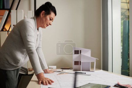 Photo for Pensive mature female architect leaning on table with building blueprint and paper house - Royalty Free Image