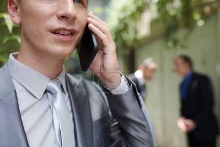 Téléchargez les photos : Entrepreneur prenant au téléphone avec un avocat lorsque ses partenaires commerciaux parlent en arrière-plan - en image libre de droit