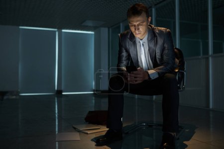 Foto de Hombre de negocios en quiebra grave sentado en la silla en el edificio de oficinas vacío y la lectura de mensajes de texto en la pantalla del teléfono inteligente - Imagen libre de derechos