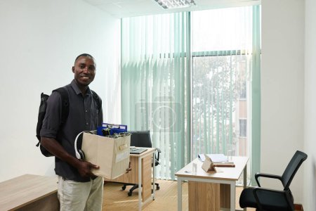 Foto de Feliz hombre negro emocionado que viene a su nuevo lugar de trabajo con caja de pertenencias - Imagen libre de derechos