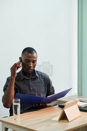 Foto de Fruncir el ceño grave Hombre de negocios negro hablando por teléfono al leer los detalles del contrato - Imagen libre de derechos