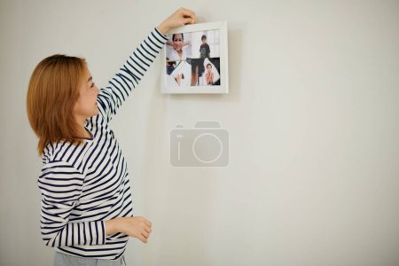 Foto de Mujer joven en camisa a rayas marco colgante con fotos en la pared en el nuevo apartamento - Imagen libre de derechos