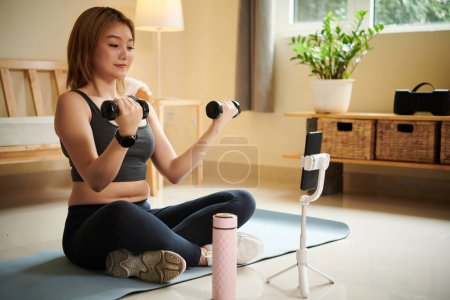 Foto de Ajuste mujer joven filmándose haciendo ejercicio con pesas para fortalecer los bíceps - Imagen libre de derechos