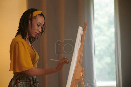 Téléchargez les photos : Créatif noir femme appréciant peinture tableau sur chevalet à la maison - en image libre de droit